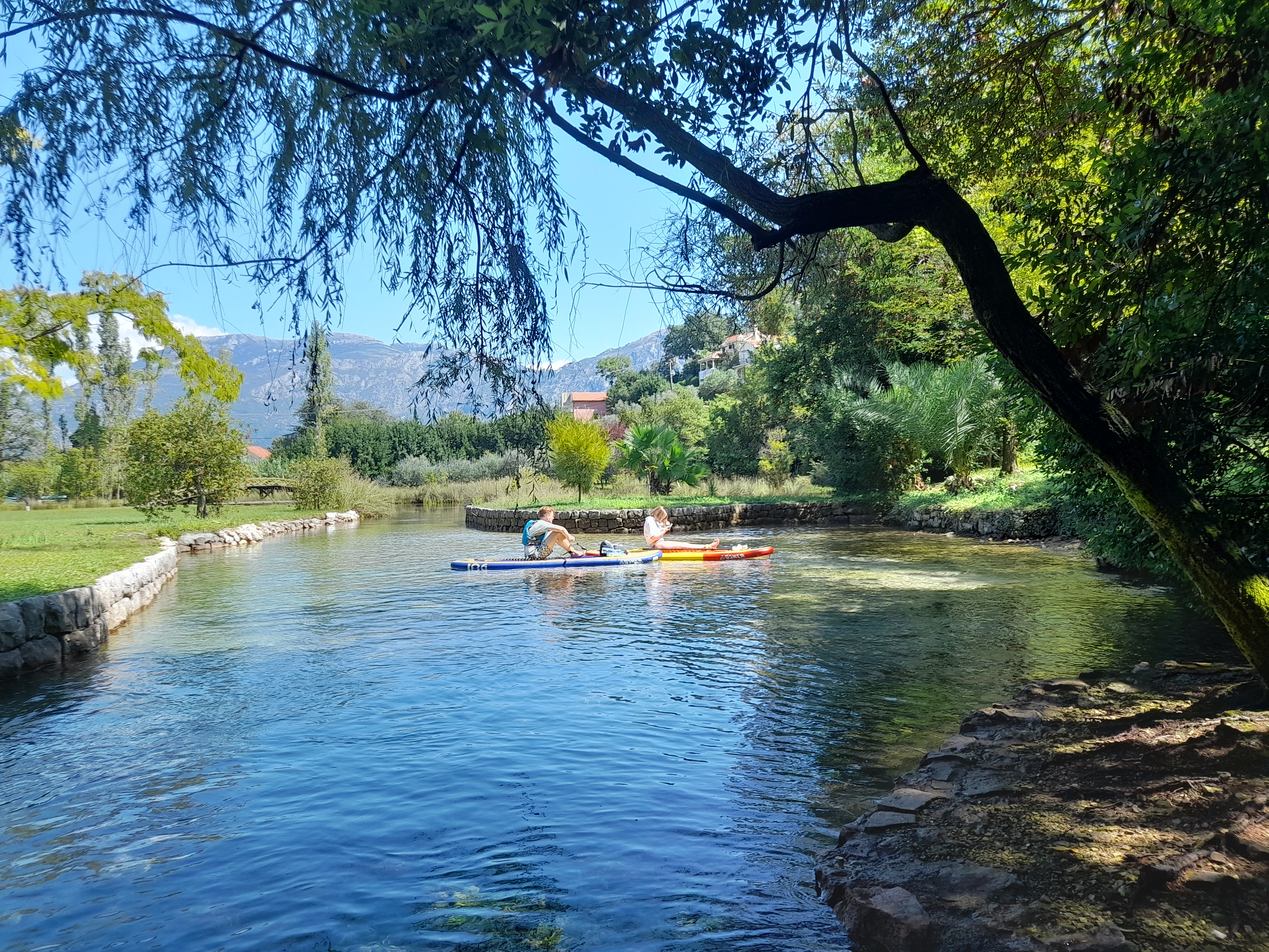 Two paddlers drifting through a lush green river channel near Perast
